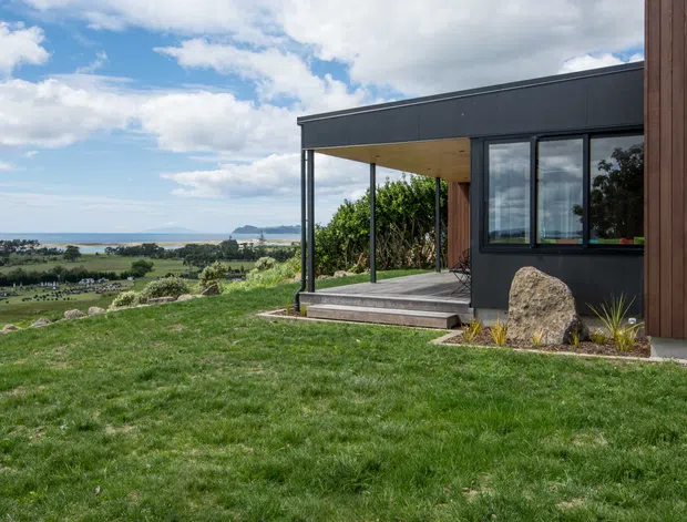 Exterior view of a contemporary single-storey house on a landscaped sloping site, with a dark flat-roofed facade, timber-clad wall, black-framed glazing, and a covered timber-lined terrace. The home overlooks open rural fields and a coastal or lakeside horizon, with a lawn, native planting, and a large natural boulder in the foreground.