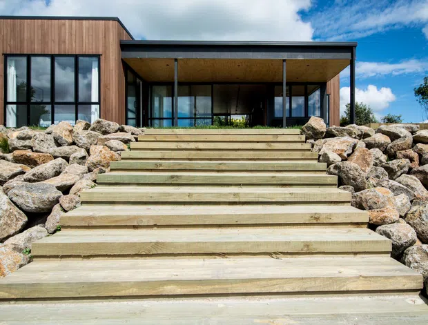 Exterior view of a contemporary single-storey house set on a rocky, sloped site. Broad timber steps lead up through landscaped rockwork to a sheltered covered entry terrace, with extensive black-framed glazing, vertical timber cladding, and a low-pitched flat roof.