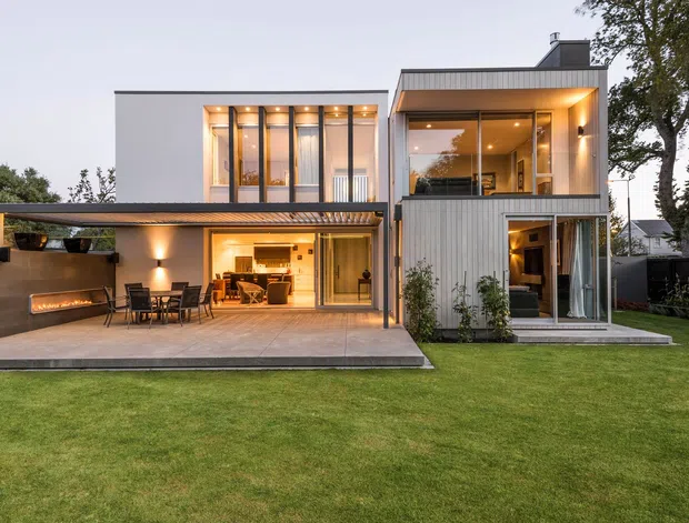 Contemporary two-storey residential home viewed from the rear garden, featuring crisp white rendered walls, vertical ribbed metal cladding, extensive floor-to-ceiling glazing, a covered outdoor entertaining area with built-in fireplace, and illuminated open-plan living spaces. A broad lawn occupies the foreground.