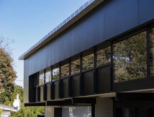 Exterior view of a contemporary dark-clad building with a long, low roofline, extensive black-framed glazing, and projecting structural elements. Mature trees and a neighbouring light-coloured roof are visible around the building.