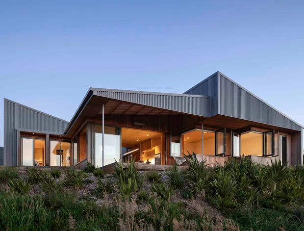 Exterior architectural photograph of a contemporary single-storey home set within landscaped coastal-style planting. The building has sharply articulated pitched and gabled roof forms, dark vertically ribbed metal cladding, extensive floor-to-ceiling glazing, and a broad timber-lined covered outdoor living area. Warm interior lighting is visible through the windows and open edges of the sheltered terrace.