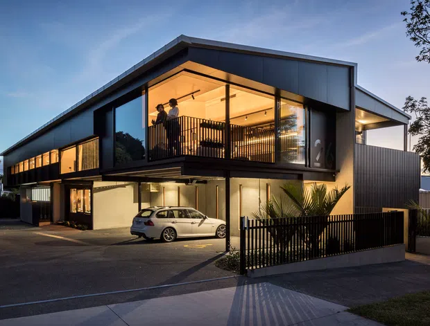 Exterior dusk view of a contemporary two-storey residence with a strong cantilevered upper volume, flat rooflines, dark metal cladding, extensive glazing, and warm interior illumination. A covered carport and driveway occupy the ground level, while a sheltered upper balcony with black vertical balustrading is prominent. Low fencing and tropical-style planting frame the street frontage.