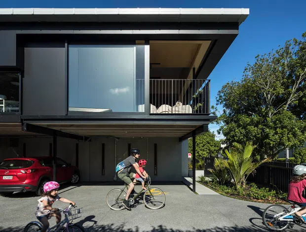 Exterior view of a contemporary two-storey residence with a projecting upper level, dark cladding, broad glazing, and a covered carport or undercroft. The upper balcony has a slim dark metal balustrade and a recessed ceiling, while landscaped planting and a paved forecourt sit alongside the building. Cyclists and parked cars are present in the foreground.