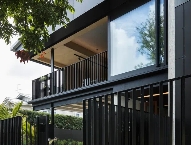 A close exterior view of a contemporary multi-level urban residence. The facade combines dark vertical metal screening and railings, exposed light-grey concrete, large black-framed glazing, and a projecting upper balcony. Tropical foliage partially frames the building, while a neighbouring suburban house and landscaped frontage are visible beyond.