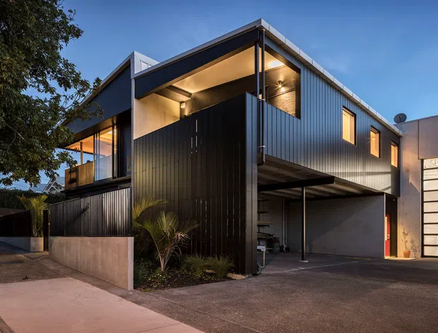 Exterior architectural photograph of a contemporary multi-level urban residence at dusk. The building features dark vertically ribbed cladding, sharp box-like forms, recessed warm lighting, upper-level glazing, a projecting canopy, and an integrated covered parking area. A large glazed garage door is visible on the right, with a landscaped edge and paved driveway in the foreground.