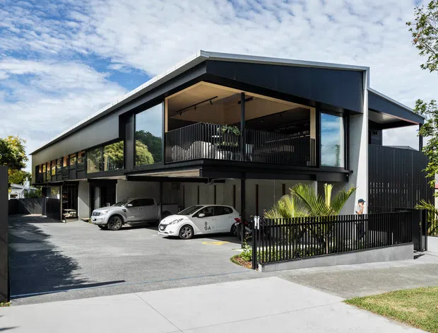 A modern multi-storey building viewed from the street, featuring a bold dark metal-clad facade, low-pitched roof forms, extensive glazing, and a large covered upper-level terrace or balcony enclosed with vertical metal balustrades. The ground level includes an open parking area and driveway, with landscaped planting and a pedestrian footpath in front.