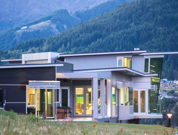 Exterior view of a contemporary multi-level house set against a steep forested mountain landscape. The home features a flat, asymmetrical roofline, clean horizontal volumes, dark and light cladding, extensive glazing, projecting balconies or canopies, and illuminated interiors visible through the windows and doors.
