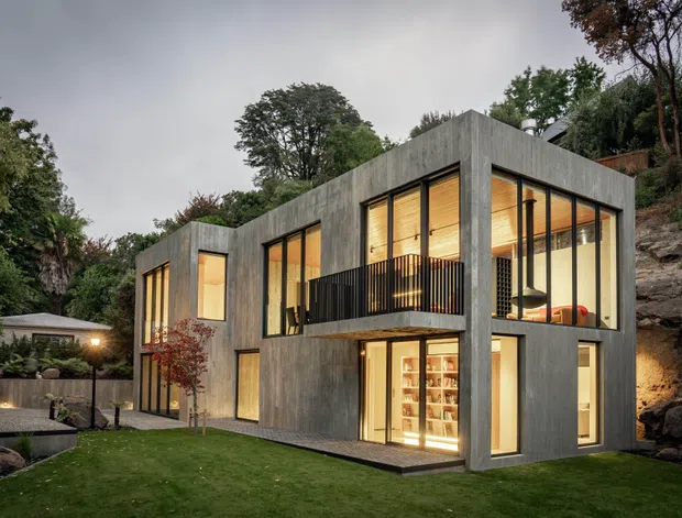 A contemporary two-storey concrete house set into a wooded, rocky sloping site. The home has a flat roof, expansive floor-to-ceiling glazing, warm timber-lined interiors visible through the windows, and a projecting upper-level balcony with dark metal balustrades. Landscaped lawn and garden surround the building.