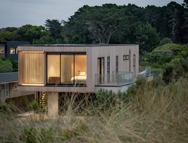 A contemporary elevated residential extension or pavilion set within dense coastal vegetation. The compact rectangular volume has grey vertical cladding, extensive dark-framed glazing, a projecting timber-slatted screen, and a glazed balcony. Warm interior lighting is visible through the windows, while tall grasses and mature trees surround the building.
