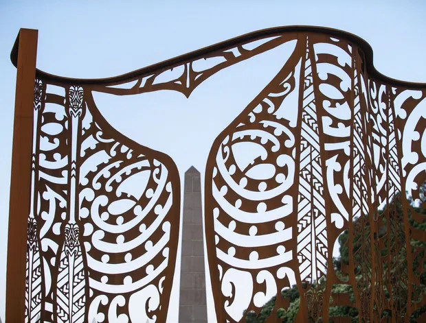 Decorative corten-steel gate or screen with ornate laser-cut botanical and scrollwork motifs, framed by curving structural members. A tall stone or concrete obelisk is visible behind it, with trees and pale sky in the background.