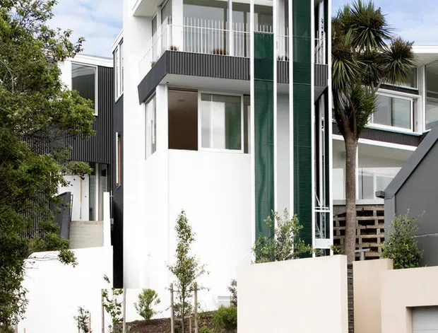 Street-facing exterior photograph of a narrow multi-storey contemporary residence. The house has a crisp white rendered facade, flat roof, projecting upper-level balcony with white vertical balustrades, dark charcoal cladding panels, and tall green screening elements. Low rendered boundary walls, landscaped planting, neighbouring homes, and a parked car are visible in the foreground.