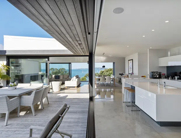 A bright, open-plan coastal home interior combining a contemporary kitchen, dining area and living space. A large white island with integrated cabinetry and bar stools anchors the kitchen, while full-height sliding glass doors open to a covered outdoor dining and living area. Polished concrete flooring, white walls, extensive glazing and timber-lined ceiling panels create a seamless indoor-outdoor connection, with ocean views visible beyond.