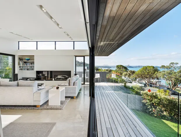 Bright contemporary open-plan living room opening onto a large timber-decked balcony or terrace. The interior features white upholstered sofas, a built-in media wall, polished concrete flooring, expansive black-framed glazing and a white ceiling with recessed linear lighting. A broad timber soffit or overhang continues above the terrace, with frameless glass balustrading and views toward a leafy coastal or waterfront setting.