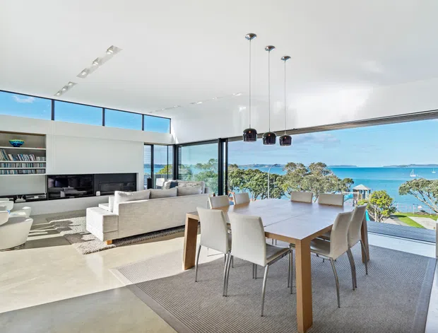Bright open-plan living and dining area with floor-to-ceiling glazing overlooking a coastal waterfront scene. The room features a pale polished concrete floor, white walls and ceiling, a built-in media and shelving unit, a large light upholstered sofa, contemporary dining table and chairs, and three black pendant lights. High clerestory glazing and expansive sliding glass doors create an airy, modern interior.