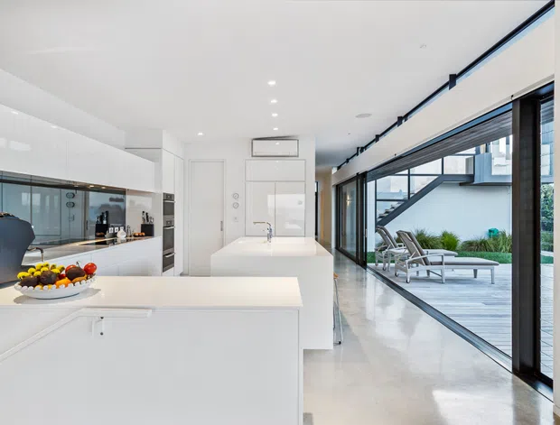 Bright, minimalist contemporary kitchen with extensive glossy white built-in cabinetry, a large white island and integrated appliances. The open-plan space extends toward full-height black-framed glazing and a timber outdoor deck with poolside loungers. Polished light flooring, recessed ceiling lights and clean linear detailing create an airy, refined interior.