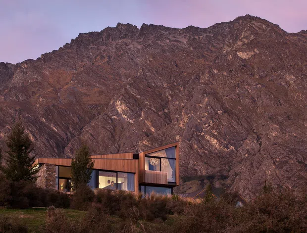 A contemporary multi-level house set against a dramatic rugged mountain backdrop. The home features dark timber or metal cladding, extensive glazing, projecting box-like volumes, and a strong angular roofline. The scene is photographed at dusk with muted purple and warm brown tones, surrounded by trees and natural landscaping.