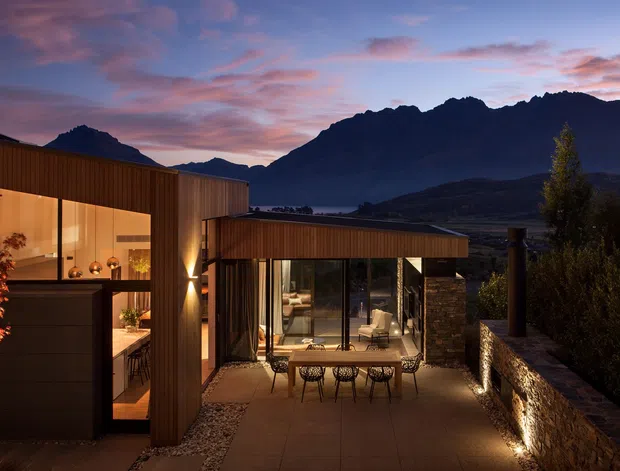 A contemporary multi-level residence at dusk, set against dramatic mountain peaks. The home features warm timber-toned vertical cladding, extensive floor-to-ceiling glazing, a sheltered outdoor dining terrace, and a low stone retaining wall with integrated uplighting. Interior furnishings and a kitchen/dining area are visible through the glass, while landscaped planting frames the approach.
