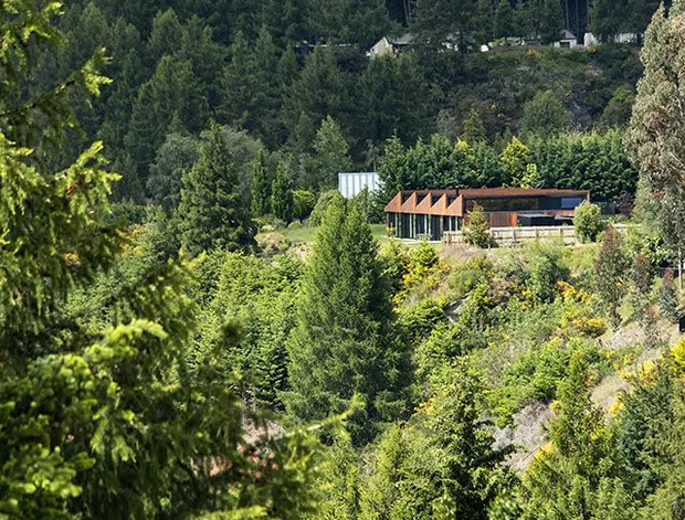 A distant exterior view of a low, elongated building set within dense, heavily planted mountainous terrain. The building has a warm reddish-brown pitched roof with repeated gabled projections, dark timber-like cladding or framing, and extensive glazing along the front. Steep forested mountains rise behind it, creating a secluded alpine setting. The image is a vertical landscape photograph dominated by vegetation and natural scenery.