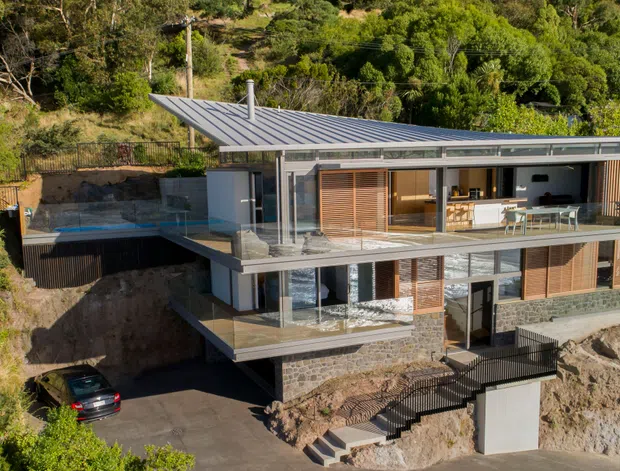A contemporary multi-level hillside residence with a low-pitched metal roof, extensive glazing, stone-clad lower walls, timber screens, and broad upper-level terraces. The home is integrated into a steep, densely vegetated site, with a swimming pool terrace, driveway, external stairs, and multiple balconies visible.