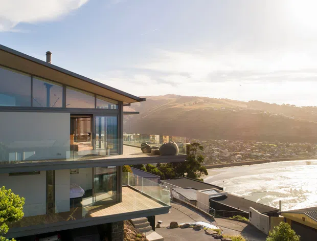 Exterior view of a contemporary multi-level hillside residence with extensive floor-to-ceiling glazing, projecting balconies, slim dark roof planes, and glass balustrades. The house overlooks a sunlit coastal or waterfront settlement and bay, with stepped terrain and landscaped surroundings visible around the property.