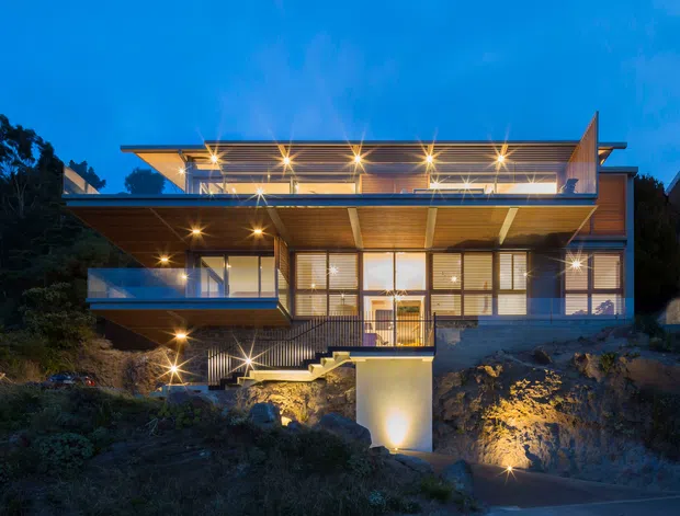 Twilight exterior photograph of a contemporary multi-level residence built into a sloping, rocky site. The house features extensive glazing, projecting cantilevered decks and roof planes, exposed timber soffits, warm recessed lighting, and a rooftop terrace or covered upper-level outdoor space. Dense vegetation and neighbouring homes frame the building.