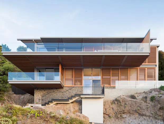 Exterior view of a contemporary multi-level house set into a sloped, landscaped site. The facade combines extensive glazing, horizontal timber cladding and louvred screens, with broad cantilevered roof planes, glass balustrades, balconies and an external stair leading to the upper level. A stone retaining wall and exposed rocky terrain are visible at the base.