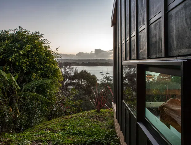 Exterior view of a dark-clad building set among dense vegetation on a sloping site, overlooking a calm waterway with boats and distant hills. The image focuses on the side facade, vertical window openings, and strong connection between the building and its verdant waterfront surroundings in soft late-day light.