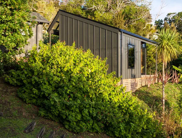 A compact modern dwelling or extension viewed from the landscaped exterior. The building has dark vertical cladding, a low-pitched roof, large narrow windows, and a light brick or masonry base, partially screened by dense planting on a sloped site. The setting appears leafy and suburban or semi-rural, with timber or masonry elements and a restrained contemporary form.