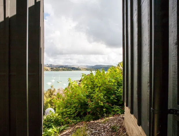 A narrow view through a partially opened dark timber-clad doorway or shutter, looking out onto a leafy garden and a calm bay or lake with small sailboats and hills beyond. The image emphasizes the relationship between the building opening and the scenic waterside landscape.