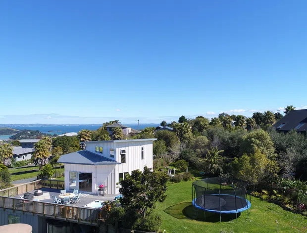 Aerial or elevated view of a contemporary two-storey residential home on a landscaped suburban coastal site. The house has light vertical cladding, a low-pitched metal roof, extensive outdoor decking and a large lawn with a trampoline. The surrounding neighbourhood and broad blue-water coastal outlook are visible beyond the property.