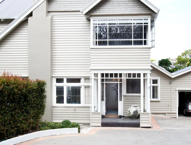 Front exterior view of a light-coloured multi-gabled house with horizontal weatherboard cladding, decorative shingle-style gable panels, a tall chimney, bay window with security grilles, covered entry porch, and attached garage. The home fronts a broad concrete driveway with hedging and small planted areas.