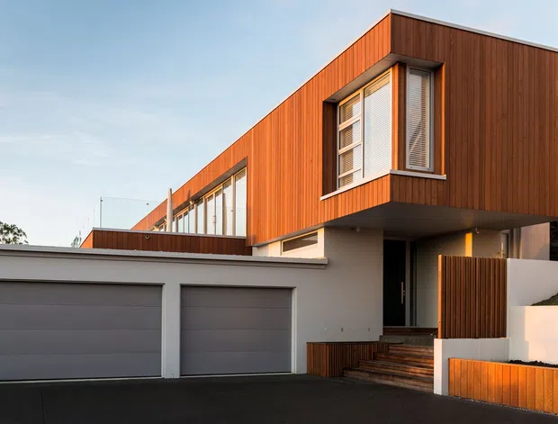 Exterior view of a contemporary multi-level residence with a strong cantilevered upper floor clad in warm vertical timber battens. The home features expansive glazing, a projecting corner window with horizontal blinds, a white rendered lower level, integrated double garage, timber entry steps and privacy fencing, and a flat roofline.