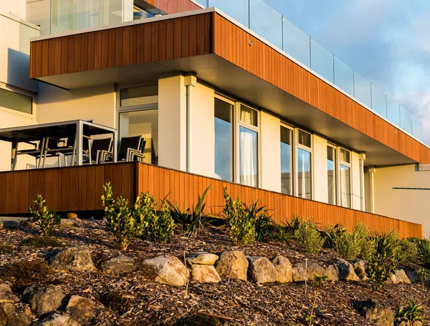 Exterior view of a contemporary multi-level home on a sloping landscaped site. The building features warm vertical timber cladding, cream rendered walls, broad projecting balconies with clear glass balustrades, and extensive floor-to-ceiling windows. A covered outdoor dining area is visible at the lower level, with rock-edged garden beds in the foreground.