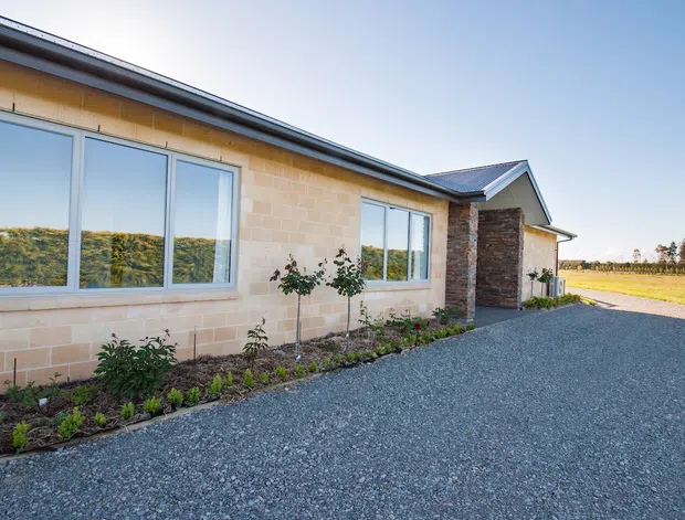 Single-storey residential building exterior with pale tan masonry walls, broad white-framed windows, a dark low-pitched roof, and a projecting entry porch faced in dark stacked stone. A gravel driveway and narrow landscaped planting bed run along the facade, with open rural fields visible beyond.