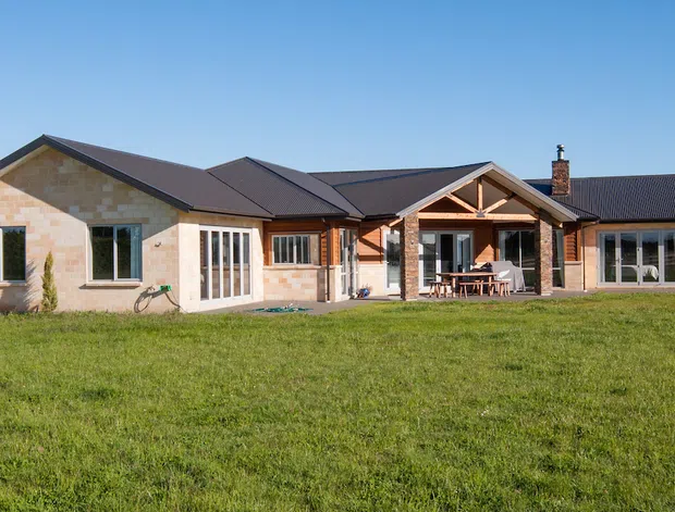 Single-storey rural home with a low-pitched multi-gabled roof, pale stone-clad and timber-clad exterior walls, dark metal roofing, exposed timber framing at the covered outdoor area, and large glazed doors and windows. The house sits in an expansive grassy rural setting with fencing and open fields.