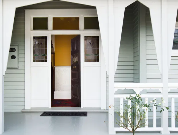 Front exterior entry porch of a traditional timber-clad house, featuring a central open doorway with decorative glazed sidelights and transom, pale weatherboard walls, white architectural trims and posts, geometric gable-like porch detailing, terracotta tiled steps, and a small landscaped garden.