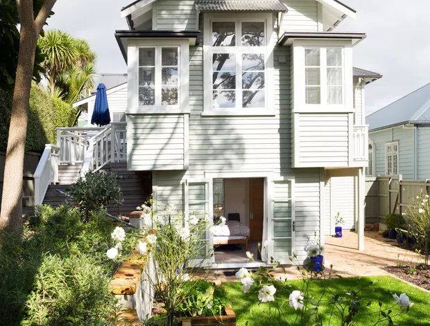 A bright exterior view of a compact multi-storey house with pale green horizontal weatherboard cladding, white trim, a prominent central gable, projecting upper-level window bays, and a small covered lower entry. The home sits within a landscaped garden with brick-edged paths, lawn, flowering plants, fencing, and mature palm-like trees. A neighbouring weatherboard house is visible to the right.