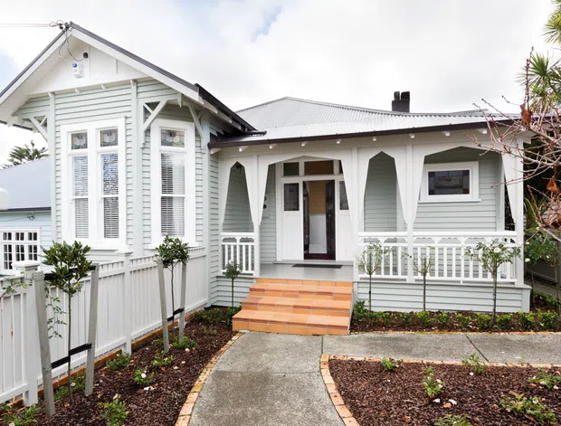 Exterior view of a single-storey light-painted weatherboard house with a prominent gabled front bay, decorative timber brackets, tall multi-pane windows, front veranda railings, a corrugated metal roof, and a landscaped path leading to the entry.