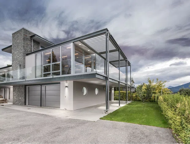 Exterior view of a contemporary multi-level house on a sloped site, featuring extensive floor-to-ceiling glazing, a projecting upper level with a covered pergola or louvred canopy, a grey rendered lower level, stone-clad vertical chimney or stair volume, balcony-like outdoor spaces, and an integrated double garage. The property is set in a landscaped suburban or semi-rural setting with mountains visible in the distance.