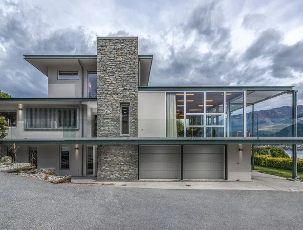 Exterior photograph of a contemporary multi-level residential house on a sloped site, featuring a flat roof, extensive glazing, a projecting upper level, a tall natural stone-clad chimney or feature wall, and an integrated double garage. A covered outdoor terrace or living area is visible at the right, with dramatic mountain and lakeside scenery beyond.