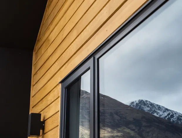 Close-up exterior architectural detail showing warm horizontal timber cladding beside a large dark-framed glazed window. A rugged, snow-dusted mountain is reflected in the glazing, with a dark soffit and wall-mounted exterior light adding contrast.