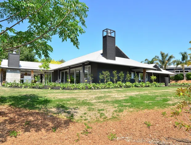 A single-storey contemporary residence set within a landscaped garden. The home has a broad low-pitched light roof, dark horizontal cladding, extensive glazing, covered verandah-like outdoor areas, and prominent rooftop chimney or ventilation structures. Mature trees and palms surround the building.