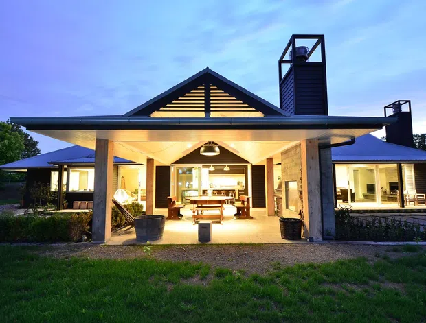 Single-storey contemporary house photographed at dusk, with a broad low-pitched roof and a prominent central gable featuring horizontal timber slats. The open central pavilion is warmly illuminated and contains an outdoor dining table, while glazed wings extend to either side. Dark metal chimney or ventilation towers rise above the roofline, and landscaped lawns and hedges surround the building.