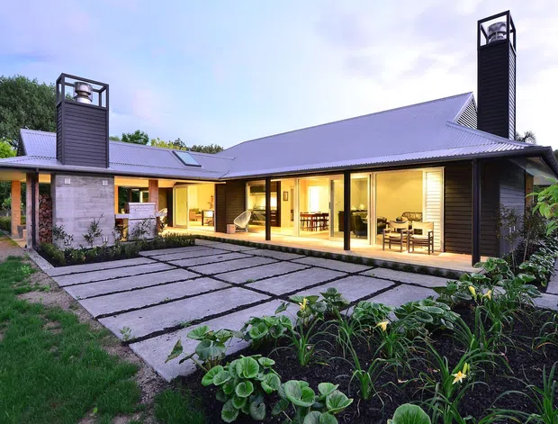 A contemporary single-storey residence viewed from the landscaped front approach at dusk. The home has a low-pitched roof, dark horizontal cladding, extensive floor-to-ceiling glazing, two tall chimney-like vertical elements, and a broad paved entry courtyard with garden beds.