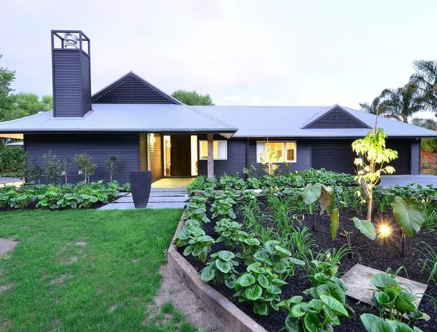 Single-storey contemporary residential house viewed from the front garden, with dark horizontal cladding, a broad low-pitched metal roof, prominent chimney, gabled roof details, warm-lit entry, and extensive landscaped garden beds.