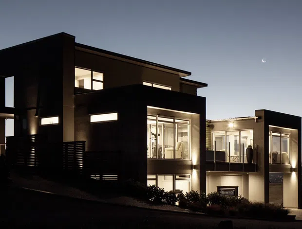 A dusk exterior photograph of a contemporary multi-level residence on a sloping site. The home has strong geometric forms, dark rendered walls, projecting box-like volumes, extensive illuminated glazing, balconies, and a flat roofline. Distant hillside lights and a crescent moon are visible in the background.
