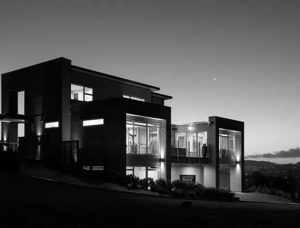 A black-and-white dusk exterior photograph of a contemporary multi-level residence on a sloping site. The home has strong rectilinear volumes, dark cladding, extensive floor-to-ceiling glazing, projecting balconies, and a small visible crescent moon above a distant illuminated landscape.