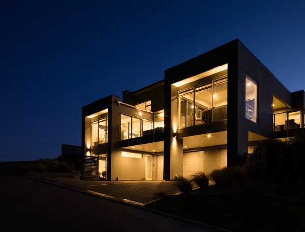 Night-time exterior photograph of a contemporary multi-level residence with strong cubic volumes, dark cladding, extensive floor-to-ceiling glazing, illuminated interiors, balconies, and recessed exterior lighting. The home sits along a paved street or driveway with minimal landscaping visible.