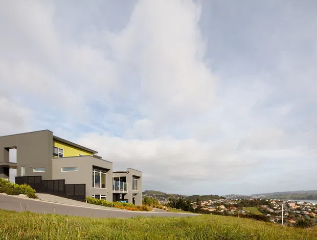 Exterior view of a contemporary multi-level residence set on a sloping site, with flat roof forms, rendered neutral walls, dark window and fence elements, and expansive glazing. The house overlooks a suburban coastal or hilly landscape under a partly cloudy sky.