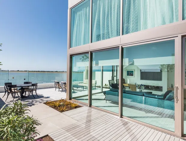 Exterior view of a contemporary waterfront residence with expansive two-storey glazing, pale framed windows, a timber deck, paved outdoor dining area, and landscaped garden beds. A bright living room with teal seating is visible through the glass, with calm water beyond the terrace.